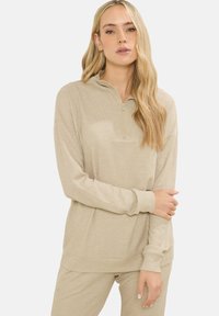 Donna con lunghi capelli biondi, vestita con un maglione beige a coste con zip e pantaloni abbinati, in piedi davanti a uno sfondo bianco semplice.