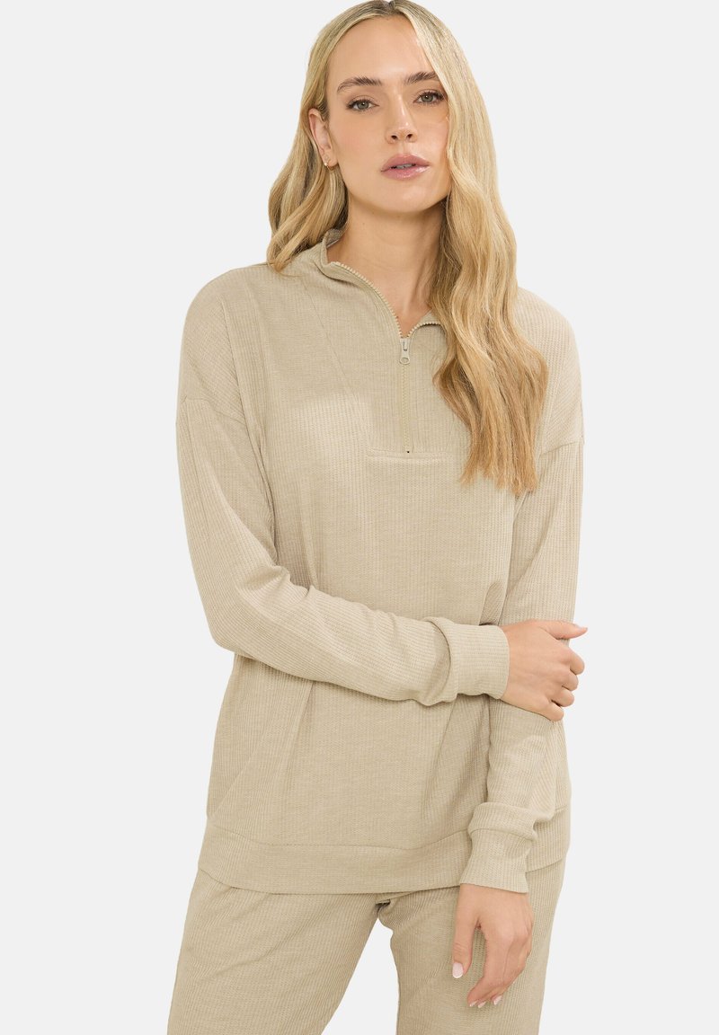 Donna con lunghi capelli biondi, vestita con un maglione beige a coste con zip e pantaloni abbinati, in piedi davanti a uno sfondo bianco semplice.