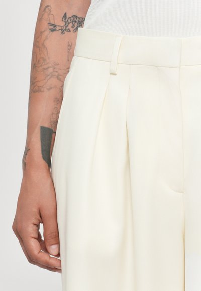 Cream-colored pleated trousers with a smooth texture, featuring a fitted waistband and side pockets, visible on a partially tattooed arm.