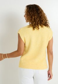 Suéter amarillo de manga corta con un acabado suave y texturizado. Presenta un cuello redondo y un dobladillo recto. Se lleva con pantalones blancos.
