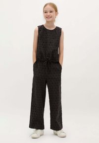 Zwarte mouwloze jumpsuit van textuurstof, met verhoogde stippen, wijde benen en een trekkoord taille; gecombineerd met witte sneakers.