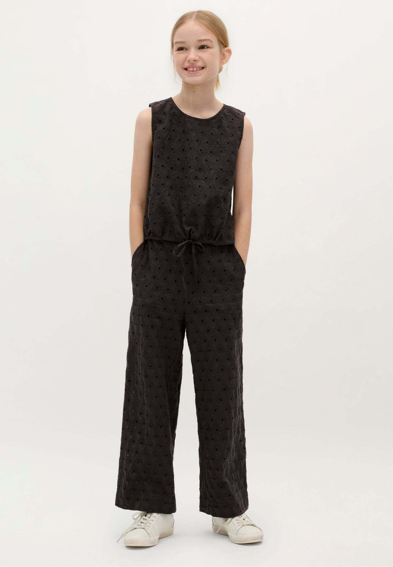 Zwarte mouwloze jumpsuit van textuurstof, met verhoogde stippen, wijde benen en een trekkoord taille; gecombineerd met witte sneakers.