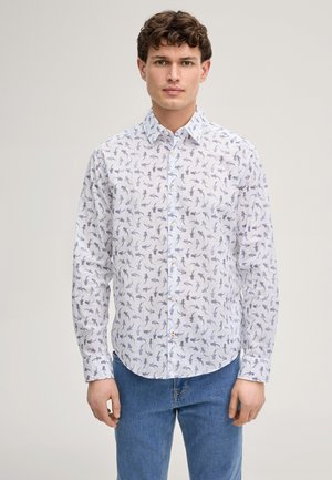 Uomo che indossa una camicia bianca a maniche lunghe con piccoli motivi a foglia blu scuro e jeans blu, rivolto in avanti contro uno sfondo semplice.