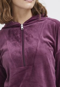 Paarse fluwelen hoodie met een ritssluiting aan de voorkant, een capuchon en een grote zak. Zachte textuur met een gladde afwerking en een relaxte pasvorm.