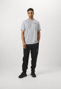 Lichtgrijs t-shirt met een kleine grafische print. Zwarte sweatpants met een logo in een donkerdere tint. Zwarte sportschoenen maken de outfit compleet.