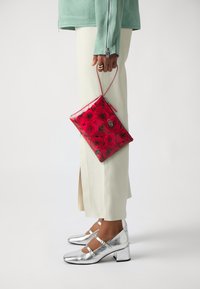 Clutch con motivo floreale rosso realizzato in materiale lucido, tenuto con una sottile tracolla. Scarpe con tacco metallico argentato che si abbinano a pantaloni di colore chiaro.