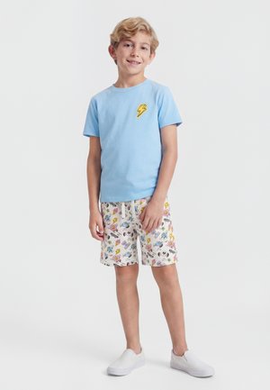 Αγόρι που φοράει ανοιχτό μπλε μπλούζα με κίτρινο κεραυνό και λευκό σορτς με πολύχρωμα καρτούν σχέδια, στέκεται με ελαφρύ χαμόγελο.