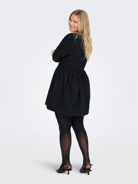 Smilende kvinne med langt blondt hår som poserer i en svart langermet kjole, svarte strømpebukser og svarte hælede sko foran en ensfarget bakgrunn.