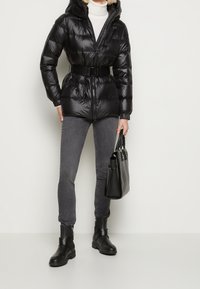 Sort dunjakke med hætte, lynlåslukning og bælte i taljen. Kombineret med grå skinny jeans og sorte ankelstøvler, holder en sort håndtaske.