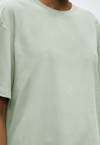 Άτομο που φοράει ένα φαρδύ, απλό ανοιχτό πράσινο κοντομάνικο T-shirt με στρογγυλή λαιμόκοψη, εμφανιζόμενο από τον λαιμό έως το μέσο του κορμού.