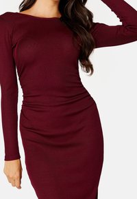 Mörkröd klänning med långa ärmar, ribbat mönster, figursydd silhuett och rynkdetaljer vid midjan, som framhäver ett mjukt, elastiskt tyg.