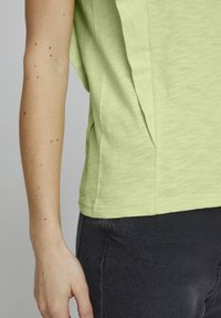 Maglietta verde chiaro con texture, dal taglio rilassato e pieghe laterali, abbinata a jeans grigi scuri. Focus sul tessuto e sul design informale.