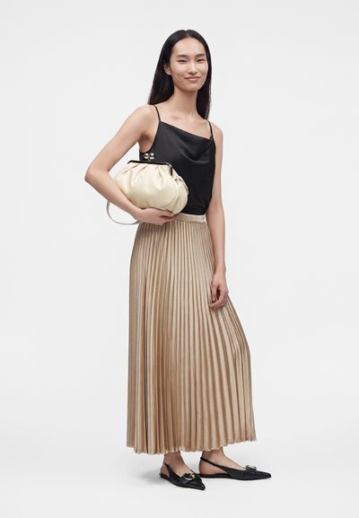 Jeune femme en haut noir sans manches et jupe plissée beige tenant une pochette crème, portant des ballerines noires à bout pointu avec une boucle.