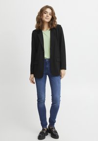 Svart skräddarsydd kavaj över en ljusgrön topp, tillsammans med blå skinnyjeans och svarta skor med kraftiga sulor. Slät tyg och figursydd design.