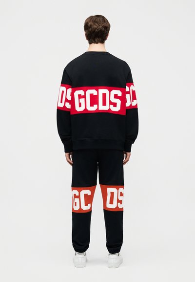 Vyras vilki juodą sportinį kostiumą su raudonomis juostelėmis ir dideliais baltais užrašais „GCDS“ ant nugaros ir kelių, stovintis veidu į priešingą pusę.