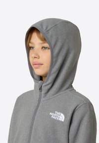 Sivá fleecová mikina na zips s kapucňou a bielym logom. Materiál má jemnú textúru a neformálny dizajn.