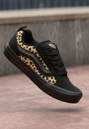 Schwarzer Sneaker mit beige-schwarzen Leopardenmuster-Seiten und schwarzer Sohle, Schnürsenkel gebunden, schwebt über einer strukturierten Bodenoberfläche.