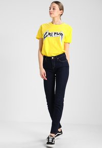 Gul grafisk t-shirt med svart text, kombinerad med slim-fit mörkblå jeans. Modellen bär svart-vita randiga sneakers.