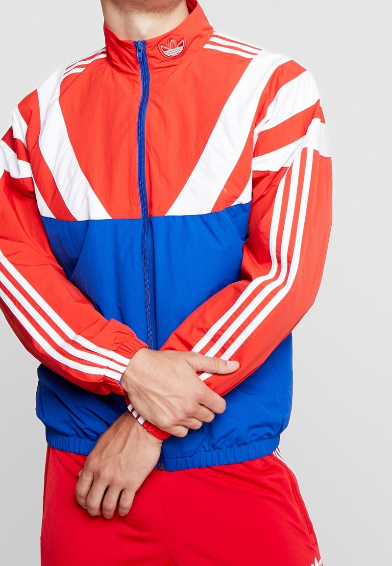 Röd, blå och vit vindjacka med hög krage, dragkedja fram och tre vita ränder på ärmarna; har en Adidas-logotyp på kragen.