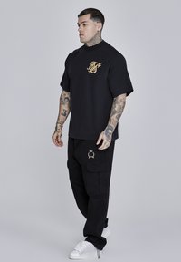 Melna pārmērīga T-kreklu ar zelta logo, kombinēts ar melnajiem cargo biksēm ar zelta emblēmu un baltajām kedām ar teksturētu detaļu.