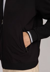 Veste noire zippée avec des poignets côtelés ornés de rayures blanches. Deux poches latérales ; tissu lisse avec une coupe décontractée.