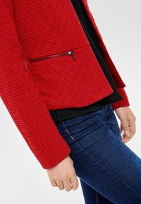 Roter strukturierter Blazer mit einer Reißverschlusstasche, kombiniert mit einem schwarzen Oberteil und blauen Jeans. Besitzt eine figurbetonte Silhouette und ein geripptes Stoffmuster.