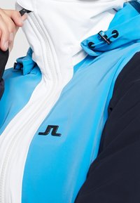 Modrá a bílá bunda se zipem vpředu, s černým logem, kontrastním lemováním a hladkým, lesklým materiálem. Obsahuje nastavitelnou kapuci.