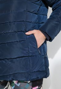 Chaqueta de plumas azul marino con diseño acolchado. Presenta un bolsillo lateral con cremallera y un ajuste cómodo. Textura suave y brillante.