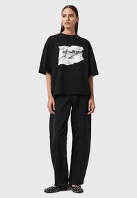 T-shirt nera oversize con una grafica bianca stropicciata recante la scritta "ALL SAINTS LONDON." Abbinata a jeans neri a gamba larga e ballerine nere con pois.