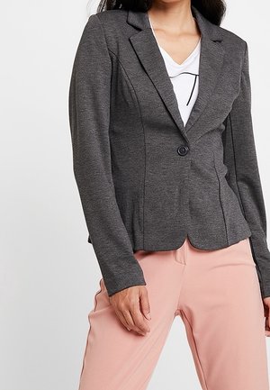 Femme portant un blazer gris foncé ajusté sur une chemise blanche au design simple, associée à un pantalon rose clair sur un fond uni.