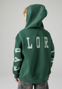 Felpa verde con una grande stampa "EXPLOR" sul retro e dettagli color crema sulle maniche. Realizzata in tessuto morbido, presenta una tasca frontale e cappuccio.