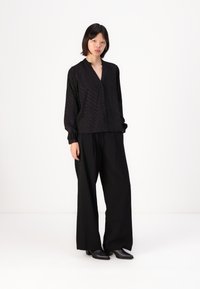 Black blouse with button closure, relaxed fit, and long sleeves. Paired with black wide-leg trousers and ankle boots. Plain background.