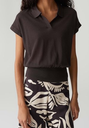 Vrouw draagt een donkerbruin overhemd met korte mouwen en een kraag, met een aangesloten tailleband, en zwart-witte broek met grote abstracte bladmotieven.