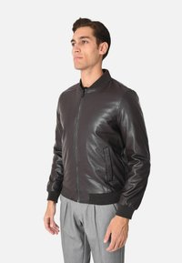 Giubbotto bomber in pelle marrone con colletto e polsini a coste, dotato di zip frontale e due tasche laterali. Texture liscia e design aderente.
