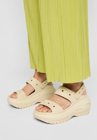 Béžové platformní sandály z gumy s otevřenou špičkou, nastavitelnými pásky a texturovanou podrážkou, nošené s plisovanými kalhotami v limetkově zelené barvě.