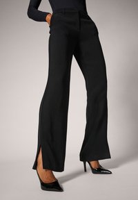 Pantalones anchos negros con aberturas laterales, diseño ajustado, textura de tela suave y pasadores para cinturón. Combinados con tacones negros puntiagudos.