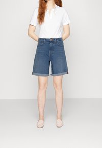 Femme portant un short en denim bleu retroussé, une chemise blanche à manches courtes avec col, et des mocassins rose clair, debout devant un fond uni.