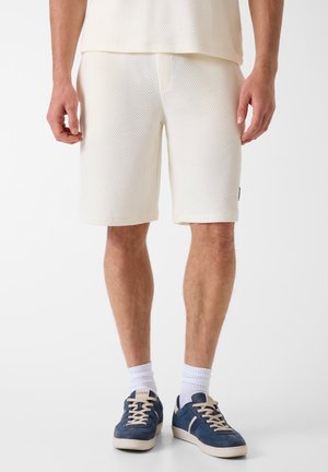 Man die een textuurrijke gebroken witte korte broek, witte sokken en marineblauwe sneakers met beige veters draagt, staande tegen een effen witte achtergrond.