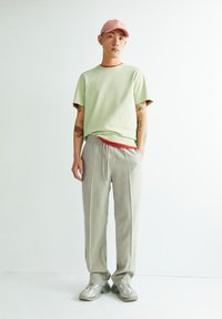 Hellgrünes Kurzarmhemd mit roten Akzenten, graue plissierte Hose und hellgraue Sneaker, getragen mit einer rosa Mütze.