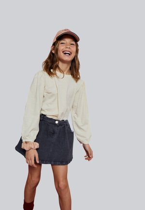 Top de manga larga color crema con mangas abullonadas, combinado con una falda de mezclilla oscura. El conjunto incluye una gorra rosa claro y calcetines rojos.