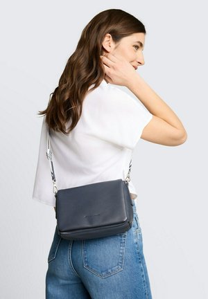 Kobieta z długimi brązowymi włosami, ubraną w białą koszulę i niebieskie dżinsy, nosząca ciemnoniebieską torbę na ramię z paskiem w paski.