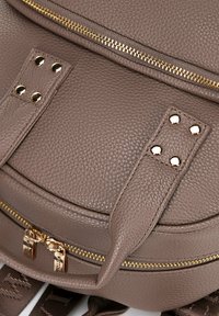 Zaino in pelle testurizzata taupe con zip dorate, quattro borchie dorate sulle cinghie e cinghie regolabili in tessuto marrone scuro.