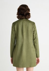 Blazer en suede vert avec des manches longues, coupe droite et détails de couture au dos. Texture lisse, longueur mi-cuisse et coupe ajustée.