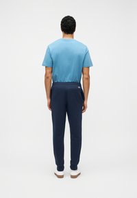 Άνδρας με σγουρά μαύρα μαλλιά που φοράει γαλάζιο μπλουζάκι, σκούρο μπλε αθλητικό παντελόνι και λευκά αθλητικά παπούτσια, στέκεται γυρισμένος με την πλάτη σε απλό φόντο.