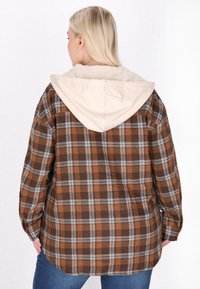 Brązowa i beżowa koszula w kratę z beżowym kapturem. Wykonana z flaneli, posiada długie rękawy, luźny krój i zaokrąglony dół.