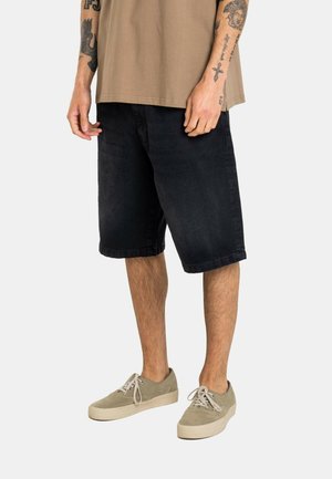 Mann trägt schwarze knielange Jeansshorts, beige Turnschuhe und ein beigefarbenes kurzärmeliges Hemd, steht mit entspannt neben dem Körper hängenden Händen.