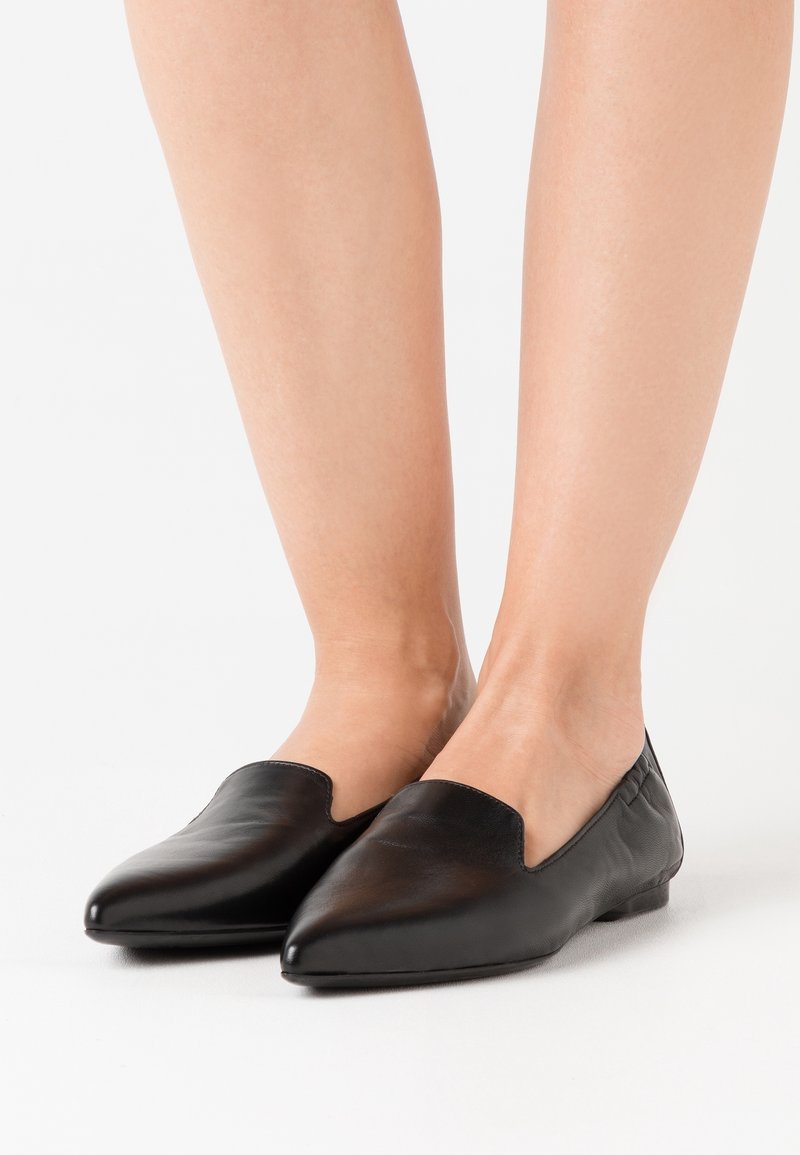 Ballerines en cuir noir à bout pointu, sans lacets, portées pieds nus sur un fond blanc uni.