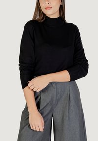 Czarny sweter z golfem z prążkowanymi mankietami, zestawiony z szarymi spodniami o wysokim stanie. Materiał wydaje się miękki i gładki.