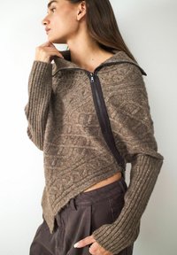 Bruine textuurtrui met een hoge kraag en asymmetrische zoom. Heeft een diagonale rits en geribbelde mouwen. In combinatie met donkere broeken.