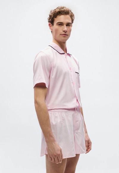 Άνδρας που φοράει ανοιχτό ροζ κοντομάνικο πουκάμισο με κουμπιά και ταιριαστά σορτς με μαύρο περίγραμμα, στέκεται μπροστά σε λευκό φόντο.